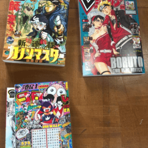 Three Japanese manga magazines with vibrant covers on a wooden surface: Boruto issue on the right featuring Naruto characters, a left issue with muscular action heroes, and a bottom-left issue with a dense collage and calendar-like page.