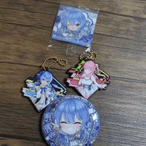 Blue-haired chibi anime character keychains and a round holographic badge on a wooden table.
