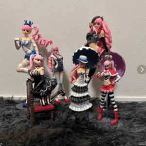 Six pink-haired fashion dolls in colorful outfits posed on a fuzzy black rug against a beige wall, with one seated on a chair and others standing, forming a playful display.