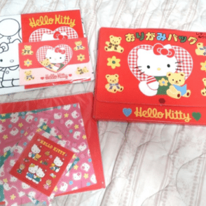 Boxed Hello Kitty memorabilia set on a quilted surface: red lunch bag with Hello Kitty graphic and yellow bears beside it, plus a matching red box and a purple/pink patterned folder set nearby.