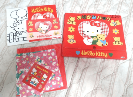 スクリーンショット 2026-06-30 111044 - Articulos Japan (post Box Japan) Boxed Hello Kitty memorabilia set on a quilted surface: red lunch bag with Hello Kitty graphic and yellow bears beside it, plus a matching red box and a purple/pink patterned folder set nearby. Articulos Japan (Post Box Japan)
