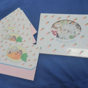 Two pastel greeting cards on blue fabric: a pink envelope with a round illustration, and a white card with a carrot pattern and an oval window showing a drawing.
