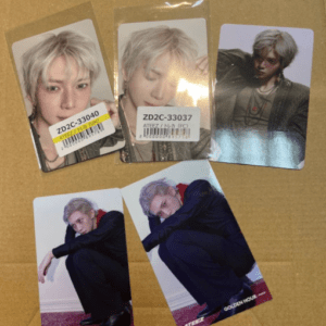 Five glossy portrait cards of a person with platinum blonde hair arranged on a brown box surface; some cards are in plastic with barcode stickers.