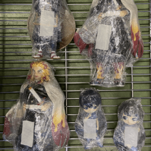 Five anime-style figurines wrapped in clear plastic sit on a metal display rack; two large figures on top, one large with a fiery cape on the bottom left, and two small blue-haired figures on the bottom right, each with a white label on their chest.