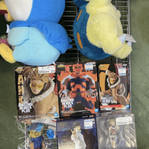 Blue plush bird and yellow-blue plush turtle on a metal rack, with anime/manga action figure boxes below (Amazing Heroes Plus, Maximatic).