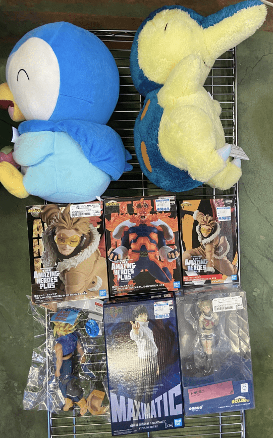 Captura De Pantalla 2026-07-02 a Las 161122 2 - Articulos Japan (post Box Japan) Blue plush bird and yellow-blue plush turtle on a metal rack, with anime/manga action figure boxes below (Amazing Heroes Plus, Maximatic). Articulos Japan (Post Box Japan)