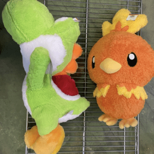 Two colorful plush toys on a metal rack: a green dinosaur-like creature with an open red mouth beside a fluffy orange chick with yellow crown-like tuft.  Articulos Japan (Post Box Japan)