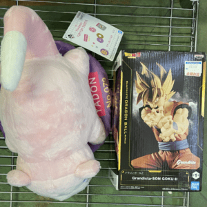 Pink plush bunny lying on a metal shelf beside a Dragon Ball Z Grandista Son Goku II figure box, with a small price tag attached.
