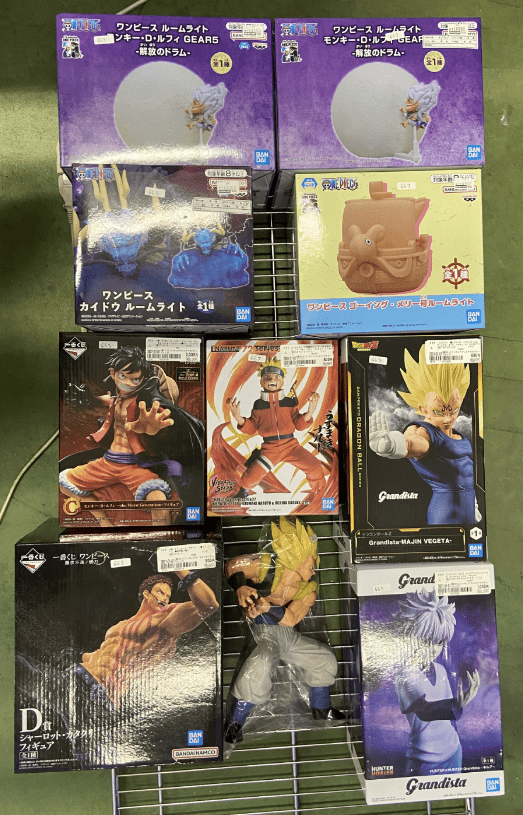 Captura De Pantalla 2026-07-03 a Las 181603 - Articulos Japan (post Box Japan) Assorted anime action-figure boxes (One Piece and Dragon Ball) arranged on a metal rack, with purple One Piece boxes on top and various Grandista boxes on the lower shelves. Articulos Japan (Post Box Japan)