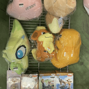 Assorted stuffed animals in clear plastic bags on a metal rack: a tall pink-lower lion, a beige hamster-like plush, a green alien with a large eye, an orange monkey, and a yellow plush cube; boxed figurines sit on the lower shelf.