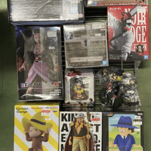 Display of boxed anime action figures on a metal grid shelf, featuring a large muscular swordsman box and various character figures.