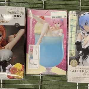 Three anime figure boxes on a metal wire rack: left box features a red-haired female figure in a white top with a flower; middle box shows a pink-haired character seated in a large blue cup; right box displays a blue-haired character in a black-and-white outfit.