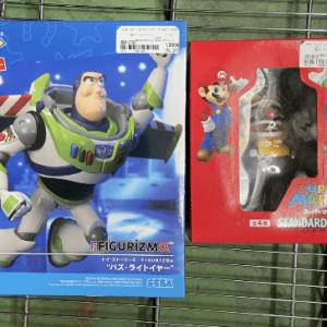 Two boxed action figures on a metal rack: Buzz Lightyear from Toy Story 5 (blue box) and a Mario figure in a red box framed by packaging glass.  Articulos Japan (Post Box Japan)