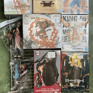Assorted anime figure boxes on a metal display rack outdoors, featuring female characters and a chibi figure with price tags visible.