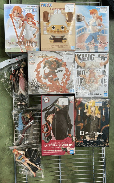 Captura De Pantalla 2026-07-19 a Las 121748 - Articulos Japan (post Box Japan) Assorted anime figure boxes on a metal display rack outdoors, featuring female characters and a chibi figure with price tags visible. Articulos Japan (Post Box Japan)