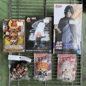 Assorted anime action figures in boxes on a metal wire rack, including chibi-style figures and a larger boxed figure (Bandai packaging).