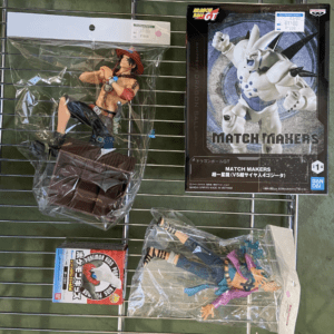 Action figures on a metal rack: a muscular character with a red hat and outstretched arms in a sealed plastic bag beside a boxed Dragon Ball GT Match Makers figure on the right position box cover shows a white armored character.