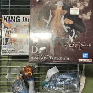 Display of anime figure merchandise on a metal shelf: a large One Piece Masterpiece box, a King of Artists poster on the left, and two smaller figures in plastic packaging (a Goku-like character and a blue mecha figure).