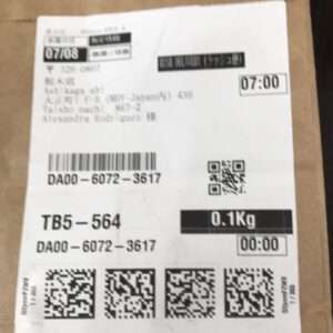White shipping label on a brown surface with multiple barcodes, QR codes, and a weight stamp reading 0.1 Kg; shows times 07:00 and 00:00 and codes like DA00-6072-3617.  Articulos Japan (Post Box Japan)