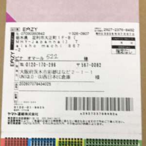 White shipping label attached to a brown box, featuring Japanese text, sender and recipient blocks, a large barcode, and color‑coded bottom strips.