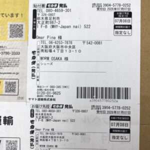 Two Japanese shipping labels with barcodes and kanji text attached to a brown cardboard box, one partially overlapping the other, on the left a yellow strip is visible.