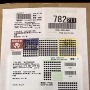 Top-down view of a cardboard parcel with multiple Japanese shipping labels, large barcodes, grid security marks, and a red handling icon.