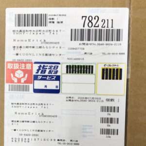 Cardboard shipping label with multiple barcodes, large tracking numbers, and Japanese text; includes red 'Handle with care' icon and blue delivery-service blocks.
