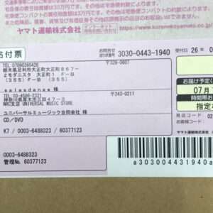Parcel shipping label in Japanese on a cardboard box, showing sender and recipient addresses, a barcode with tracking number, and a delivery date box.