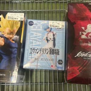 Three boxed anime figures on a metal cart: Majin Vegeta (Dragon Ball Z) left, Rei Ayanami figure in the middle, Evangelion 3.0 box on the right.
