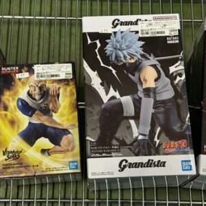 Four boxed anime figures lined up on a metal shelf: from left, a purple-haired female character figure in a dark outfit with a pink accessory box label; next, a yellow-clad muscular male figure box with blue shorts in action pose; third, a gray-clad ninja figure box featuring a character with spiky blue hair in a dynamic stance; fourth, a blond-haired character in a black outfit holding a weapon on a dark boxed figure.