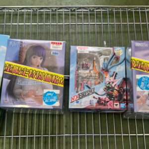 Four boxed anime figures in plastic packaging laid on a metal grid shelf, from left: a black-suited figure with a yellow hood, a purple-haired character, a bright mecha set, and a silver-haired character with blue accents.