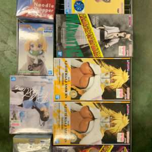 Boxed anime figures and collectibles stacked on a metal wire rack on a green floor, colorful character art visible on the boxes.