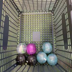 Six colorful plastic Easter eggs arranged in the bottom of a gray grid-pattern shopping basket on grass.