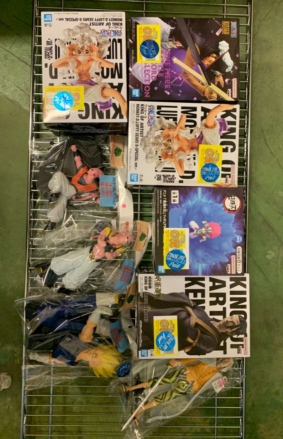 Img_2797 - Articulos Japan (post Box Japan) Metal shelving rack on a green floor holding multiple boxed anime figures and several loose figures in plastic bags; boxes feature bold graphics and character art (Dragon Ball/One Piece style). Articulos Japan (Post Box Japan)