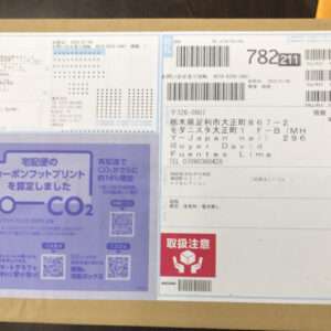 Cardboard shipping box with multiple labels: a blue address label on the right, a purple CO2 infographic on the left, and various barcodes/text in Japanese.