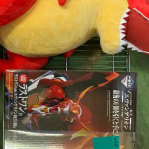 Large red-and-yellow dinosaur plush lying on its side atop a boxed action figure on a metal shelf, viewed from above.