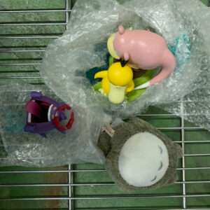 Collection of small toys and plush figures on a bubble-wrapped metal grid shelf: a pink blob creature, a yellow chick-like figure, a purple toy with a red beak, and a gray Totoro-style plush.