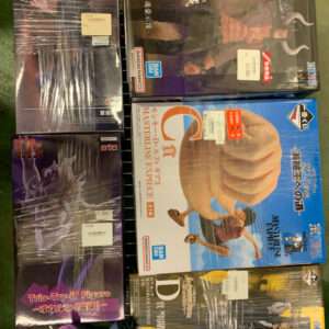 Boxed anime action figures stacked on a metal wire display rack, wrapped in plastic. The boxes are predominantly dark with blue and orange accents and Japanese text visible on the boxes and price stickers present on the packaging.],