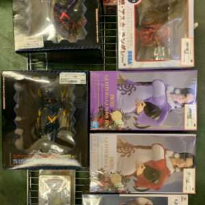 Assortment of boxed anime figures on a metal display rack, boxes with clear windows showing colorful characters and mecha. A mix of sizes and styles on a grid shelf.