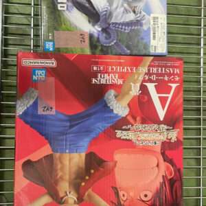 Two boxed anime figures on a metal wire rack; a large red box in front shows illustrated characters, with a smaller blue/green box behind it.  Articulos Japan (Post Box Japan)