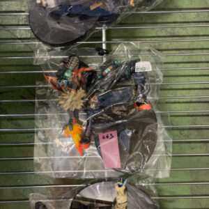 Three action figures sealed in clear bags hanging on a metal rack; one in a dark outfit with yellow grid, a spiky-haired figure, and a blue-cream armored character.