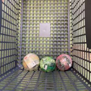 Three colorful plastic balls, yellow, green, and pink, wrapped in plastic inside a gray grid basket resting on grass