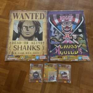 Two large illustrated posters laid on a wooden floor: a 'WANTED' poster featuring a male character labeled 'SHANKS' with 'DEAD OR ALIVE' and a bounty amount, and a colorful 'CROSS GUILD' poster with a clown and crossed swords labeled 'BUGGY'. Below are three small packaged collectible cards/jetsam.