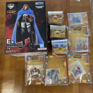 Large box showing a One Piece Masterlise Expiece figure (man in blue cape) with Bandai Namco logo; several wrapped mystery prize items beside it on a wooden floor.