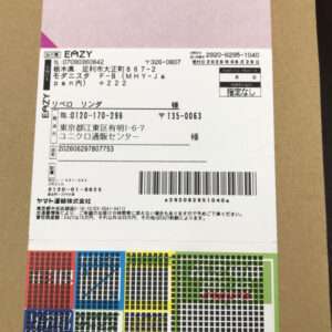 Shipping label on a cardboard box with Japanese text, a large barcode, and colorful grids along the bottom edge.