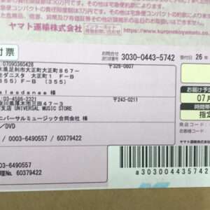 Japanese shipping label on cardboard with sender and recipient addresses, item type (CD/DVD), tracking numbers, barcode, and delivery/date fields