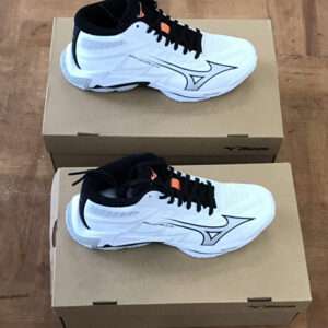 Two white Mizuno running shoes with black laces rest on top of brown shoeboxes on a wooden floor.