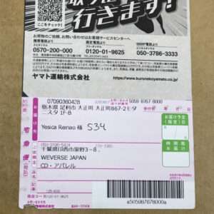 Close-up of a cardboard box with a Japanese shipping label: a purple delivery slip showing recipient name Yesica Reinao, address fields, a barcode, and a QR code; includes a URL (kuronekoyamato.co.jp) and various contact numbers.