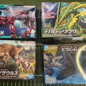 Four boxed Bandai plastic model kits on a metal wire shelf: top-left a Red Gundam box, top-right a green/yellow dragon‑mech, bottom-left a dinosaur/ Tyrannosaurus kit, bottom-right a dark blue dragon/creature kit with a circular yellow highlight.
