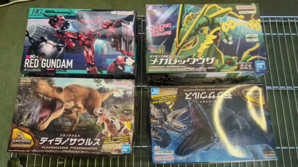 Img_e0701 - Articulos Japan (post Box Japan) Four boxed Bandai plastic model kits on a metal wire shelf: top-left a Red Gundam box, top-right a green/yellow dragon‑mech, bottom-left a dinosaur/ Tyrannosaurus kit, bottom-right a dark blue dragon/creature kit with a circular yellow highlight. Articulos Japan (Post Box Japan)
